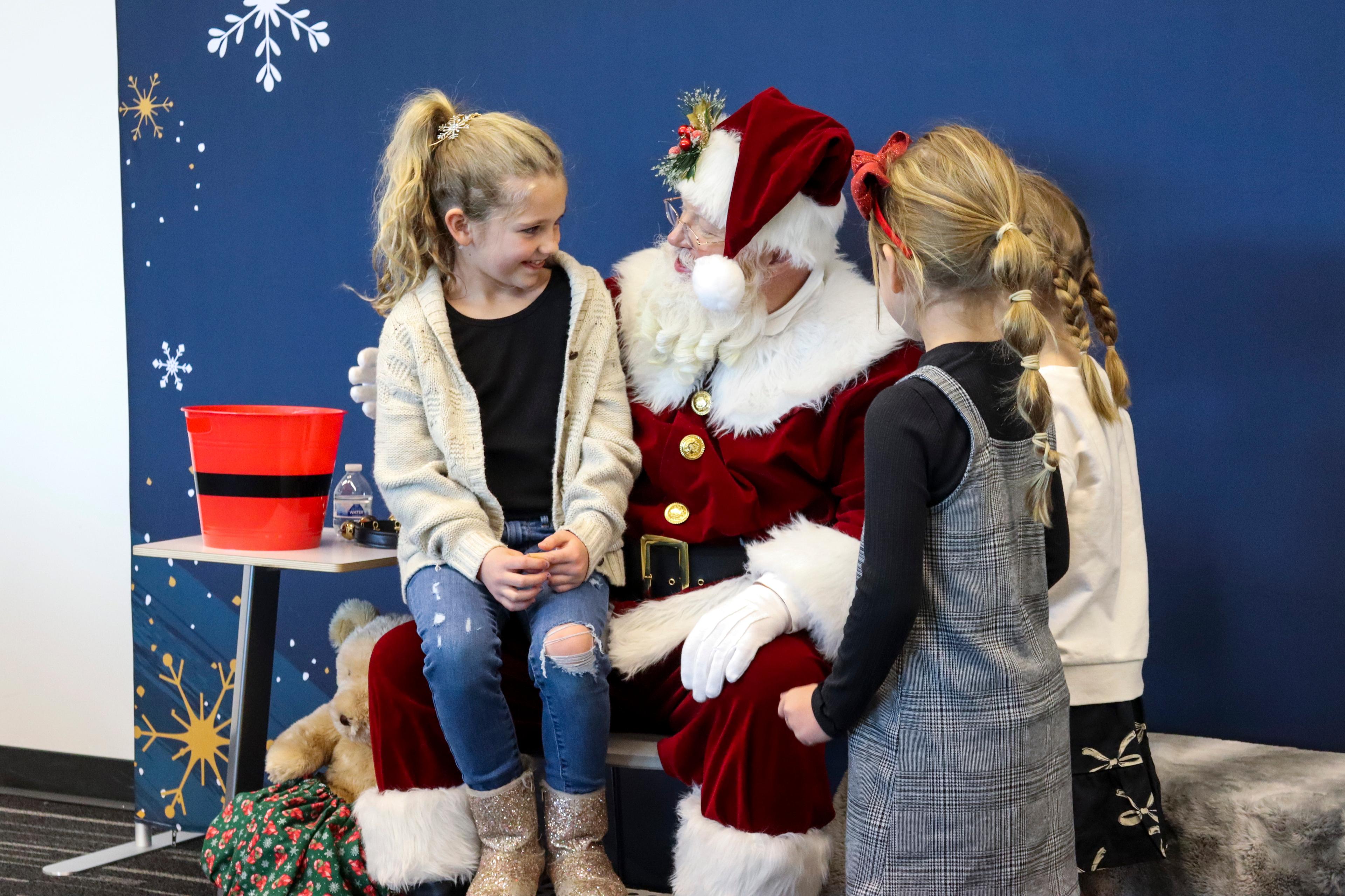 Three little girls surround a santa, with one sitting on his lap telling him what she wants for Christmas