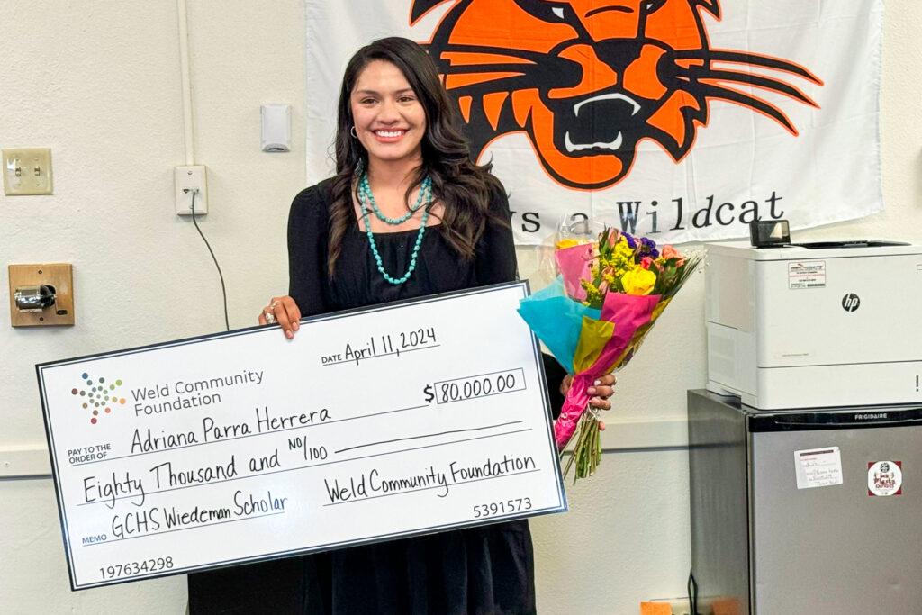 A student holds a large oversized check and a bouquet of flowers in an office.