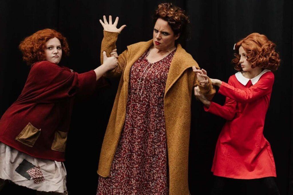 Two girls in Annie costumes tug on either side of the actress playing Miss Hannigan on stage.