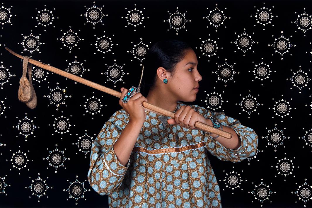 A photograph of a native person in patterned clothing holding a wooden staff with a small pouch. The background features a repeating star pattern.