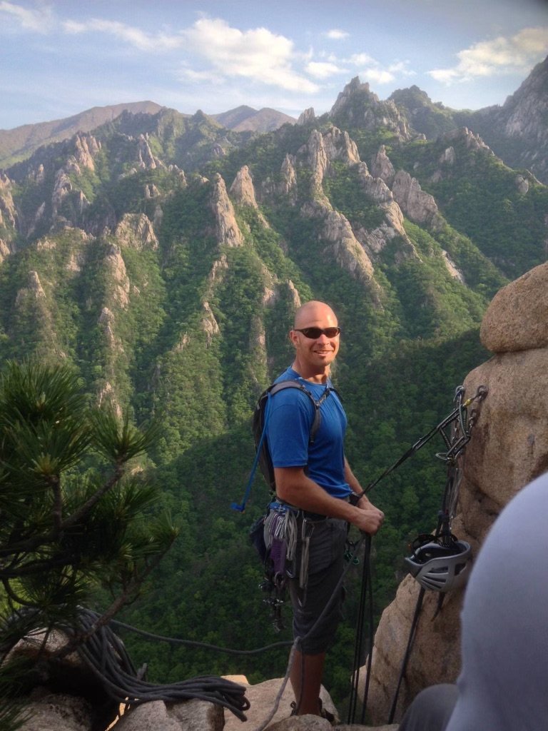 a smiling man in sunglasses and a camera slung over his shoulder is climbing a mountain. He is wearing athletic gear, and it appears he is on a very high ledge.