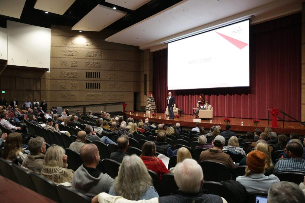 A wide shot of a theater audience watching a man speak to a microphone on stage.