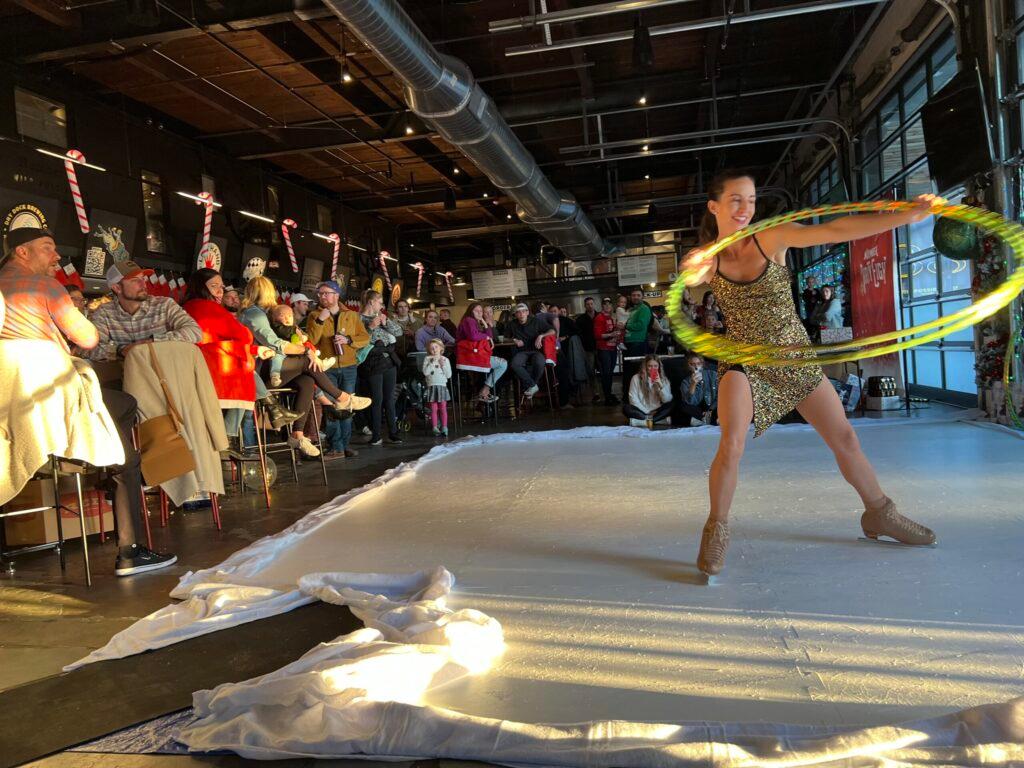 An Iceformance performer on ice skates spins a hula hoop in front of an audience in a large brewery.