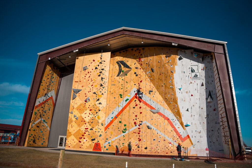 image of a climbing wall that is about 40 feet high. On the outside it's made of wood with a lot of rungs for the climbers to latch on to . It is taken outdoors and there is bright blue sky in the background