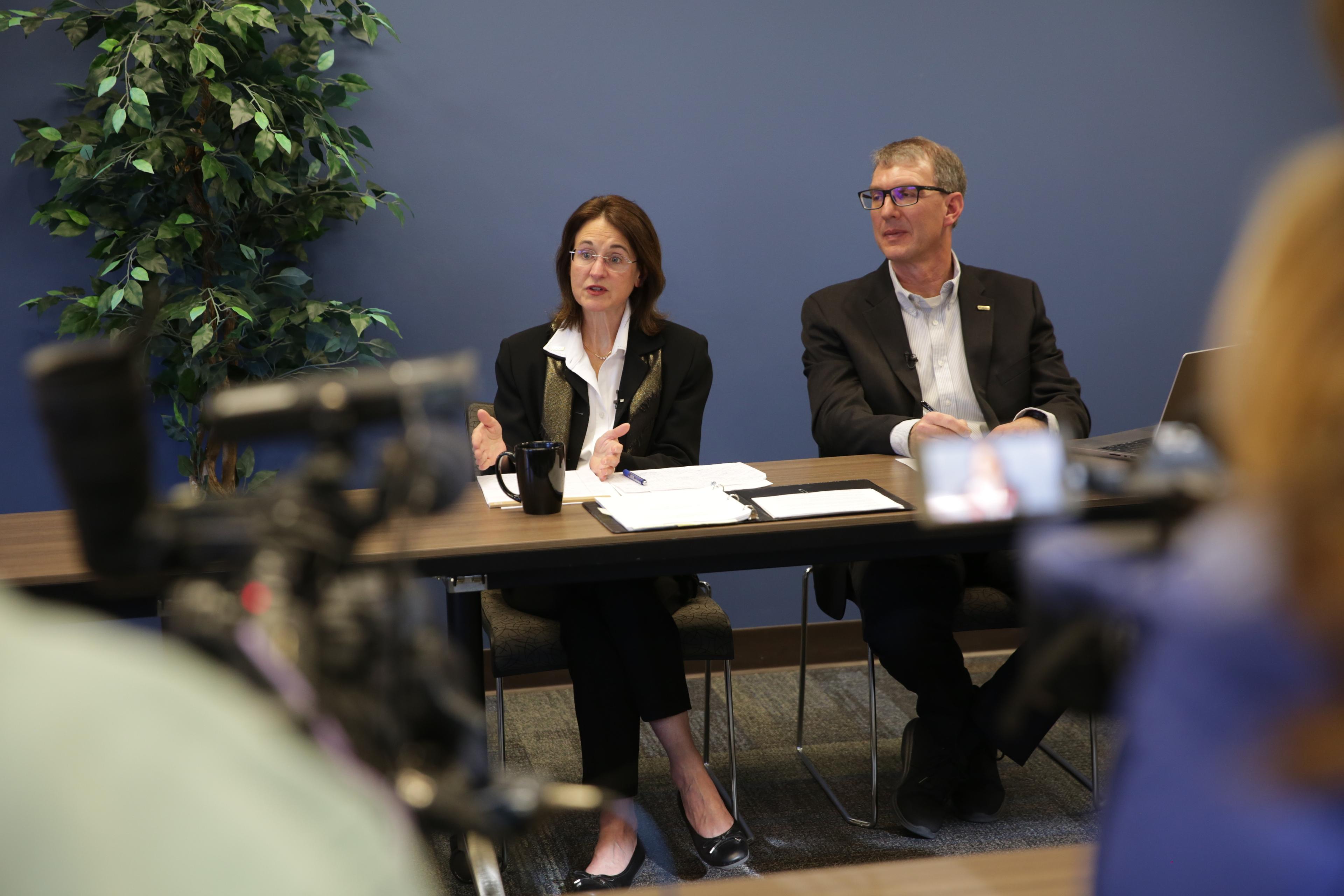 A woman and man sit at a table during a press conference. Reporters with news cameras are in the foreground.