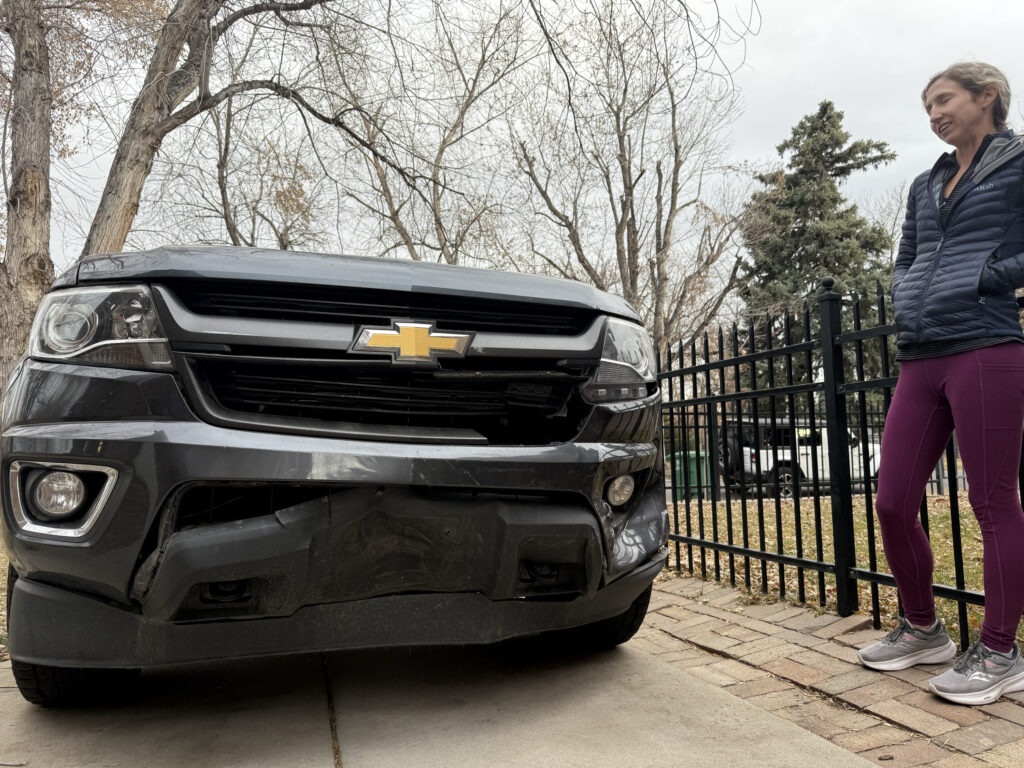 Katy Gun stands in a driveway next to a Chevrolet truck with its front bumper crumpled in, after it was hit by an uninsured driver in front of her east Denver home.