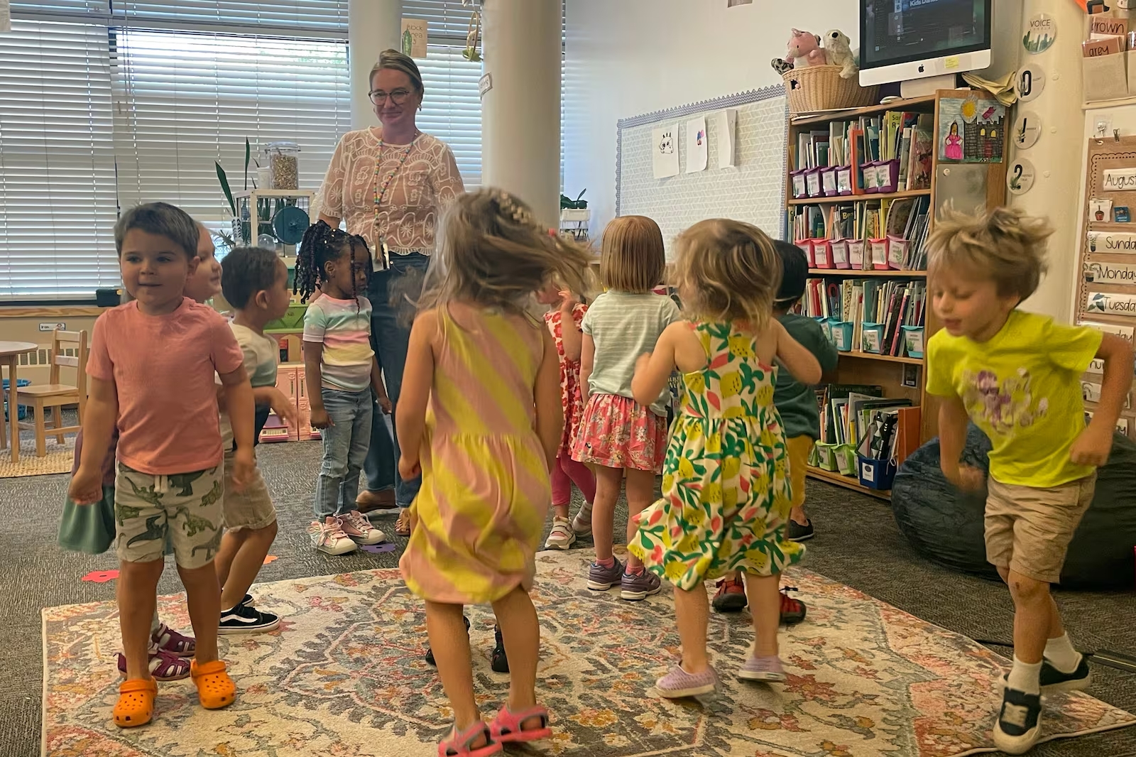 Young children playing in a circle standing and jumping around in a classroom with an adult to the side.