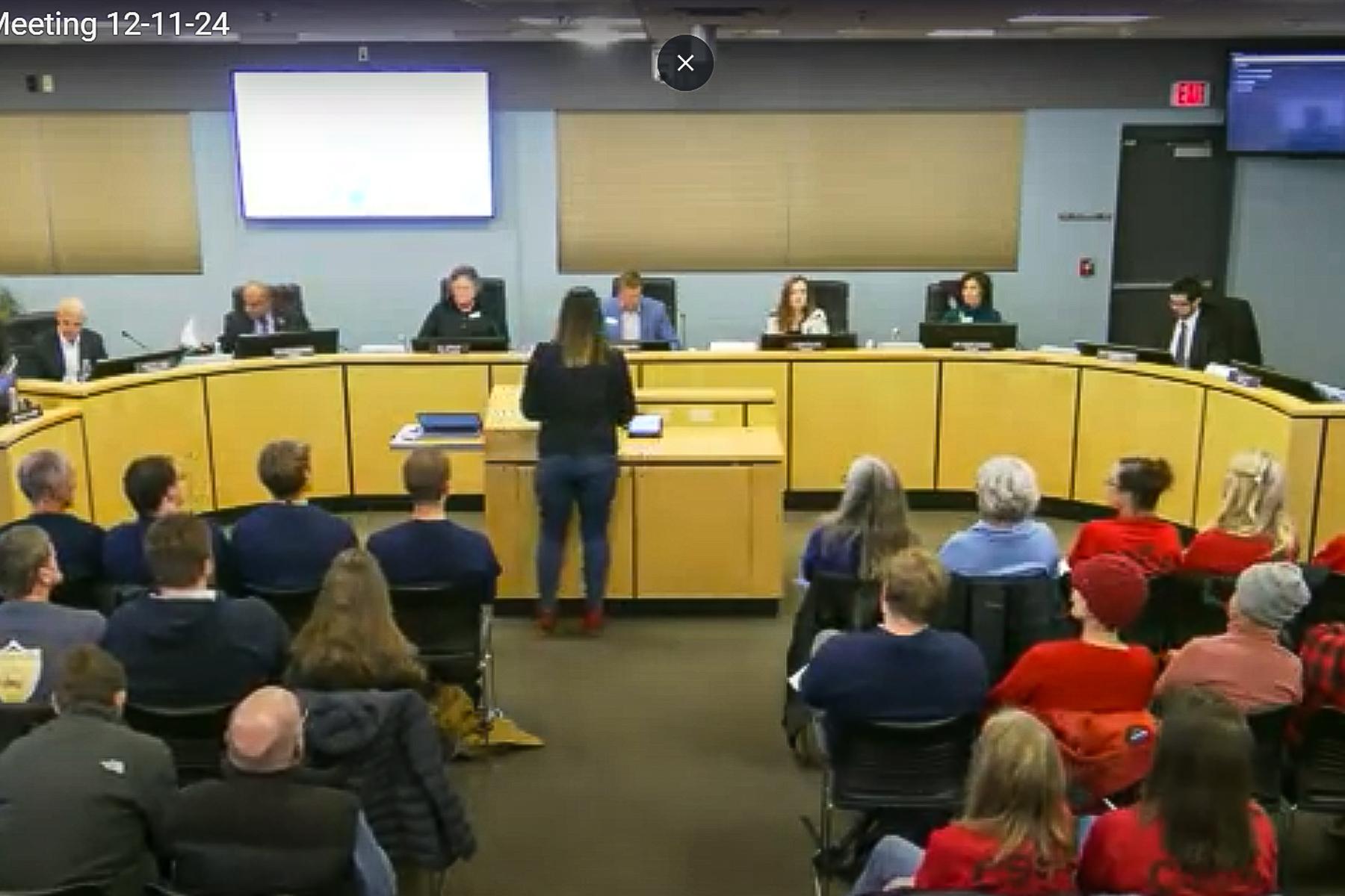 A woman stands at a lecturn with her back to the camera speaking to the seven board members seated at a rounded dais. Several rows of attendees can be seen in the foreground wearing blue an red shirts.