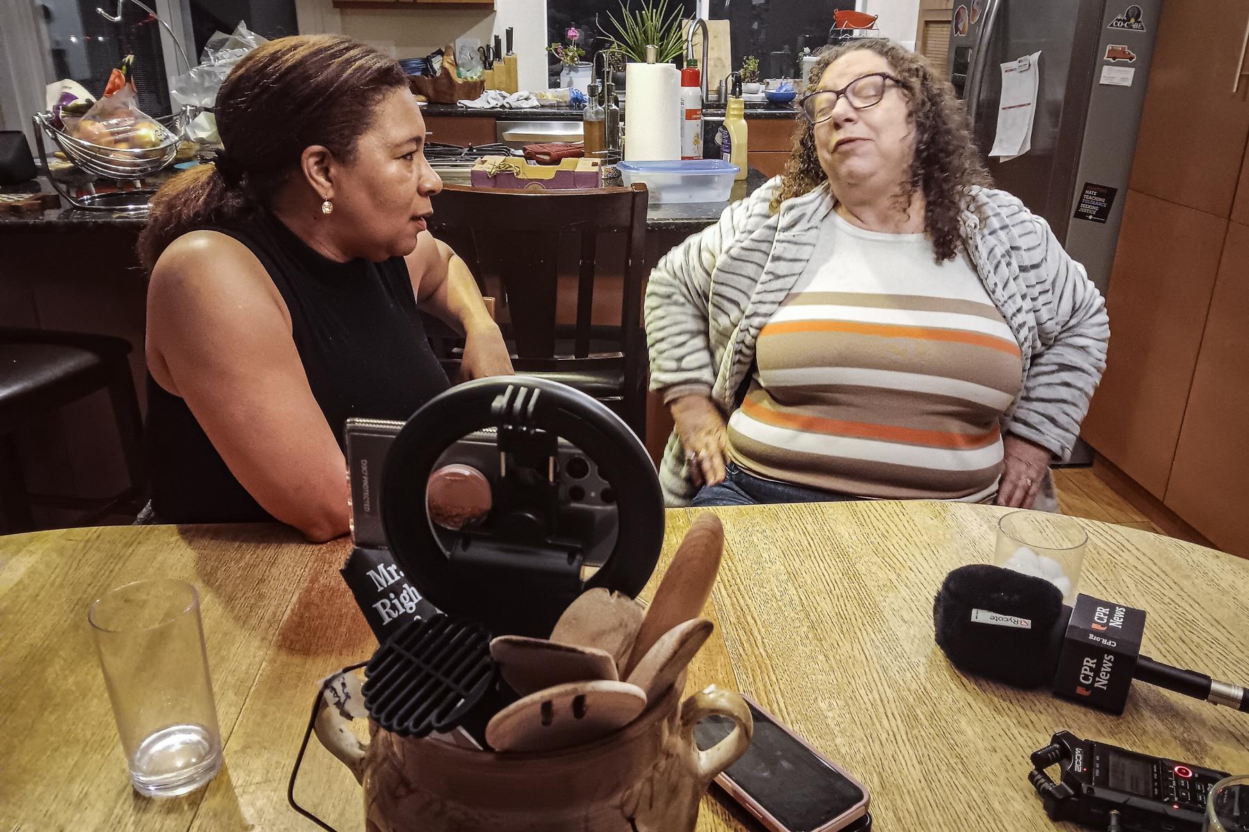 Two women sit at a table, on the left a woman looks to the woman next to her. On the right is another woman who is being interviewed. On the table in front of them are recording devices. In the background are other kitchen items.