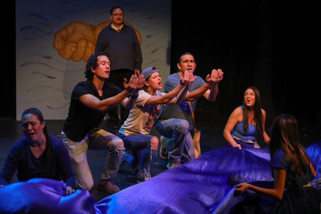 A group of people on stage perform expressively in front of a backdrop depicting a large hand and river.