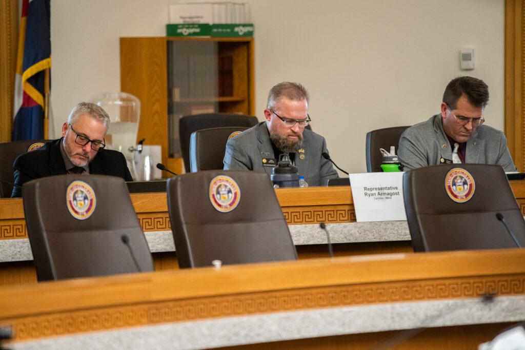 three men in suit coats sit behind a curving table, looking down