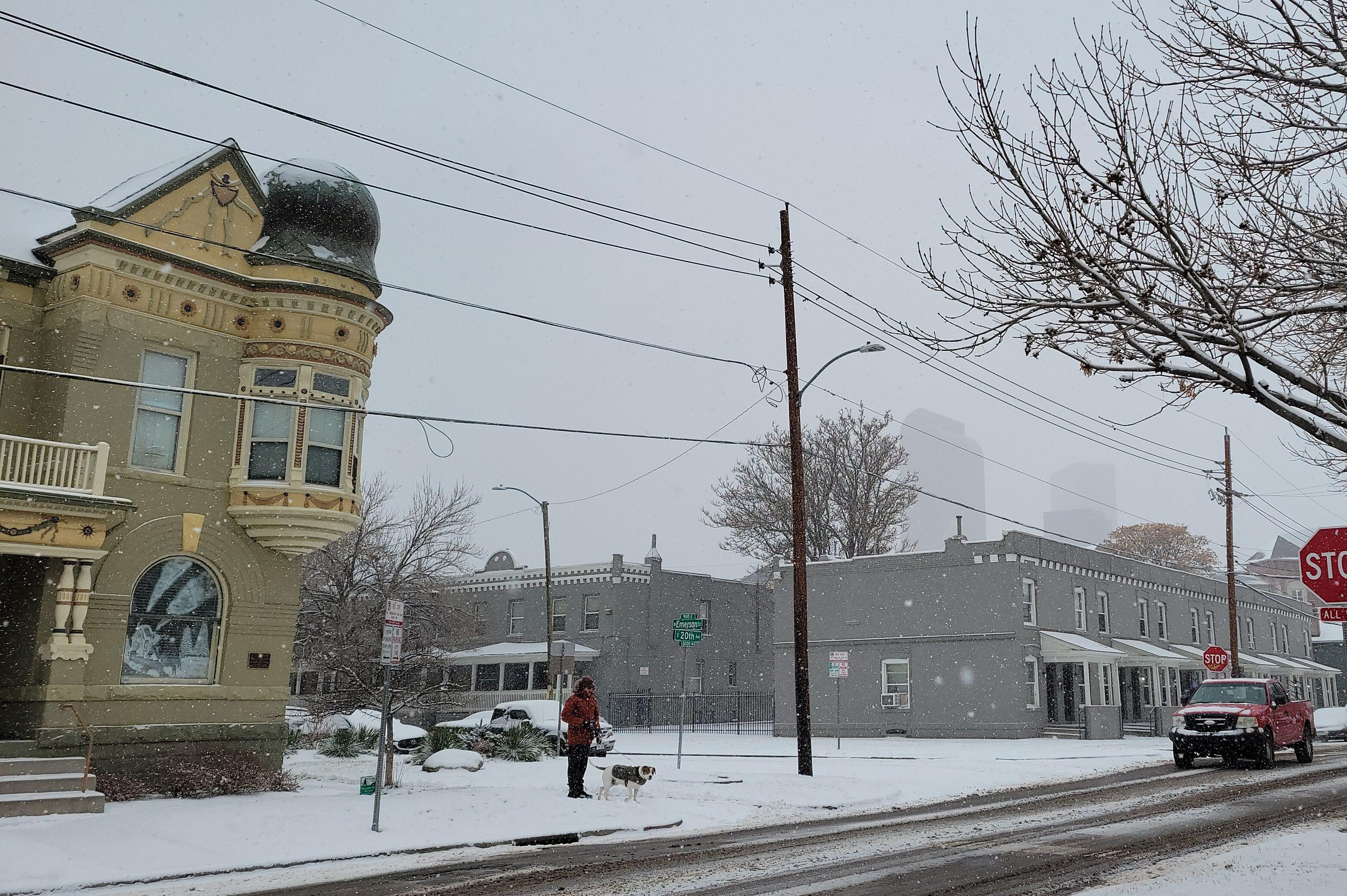 A man walks his dog in snow with a building to his right and Downtown Denver in the background shrouded in snow.