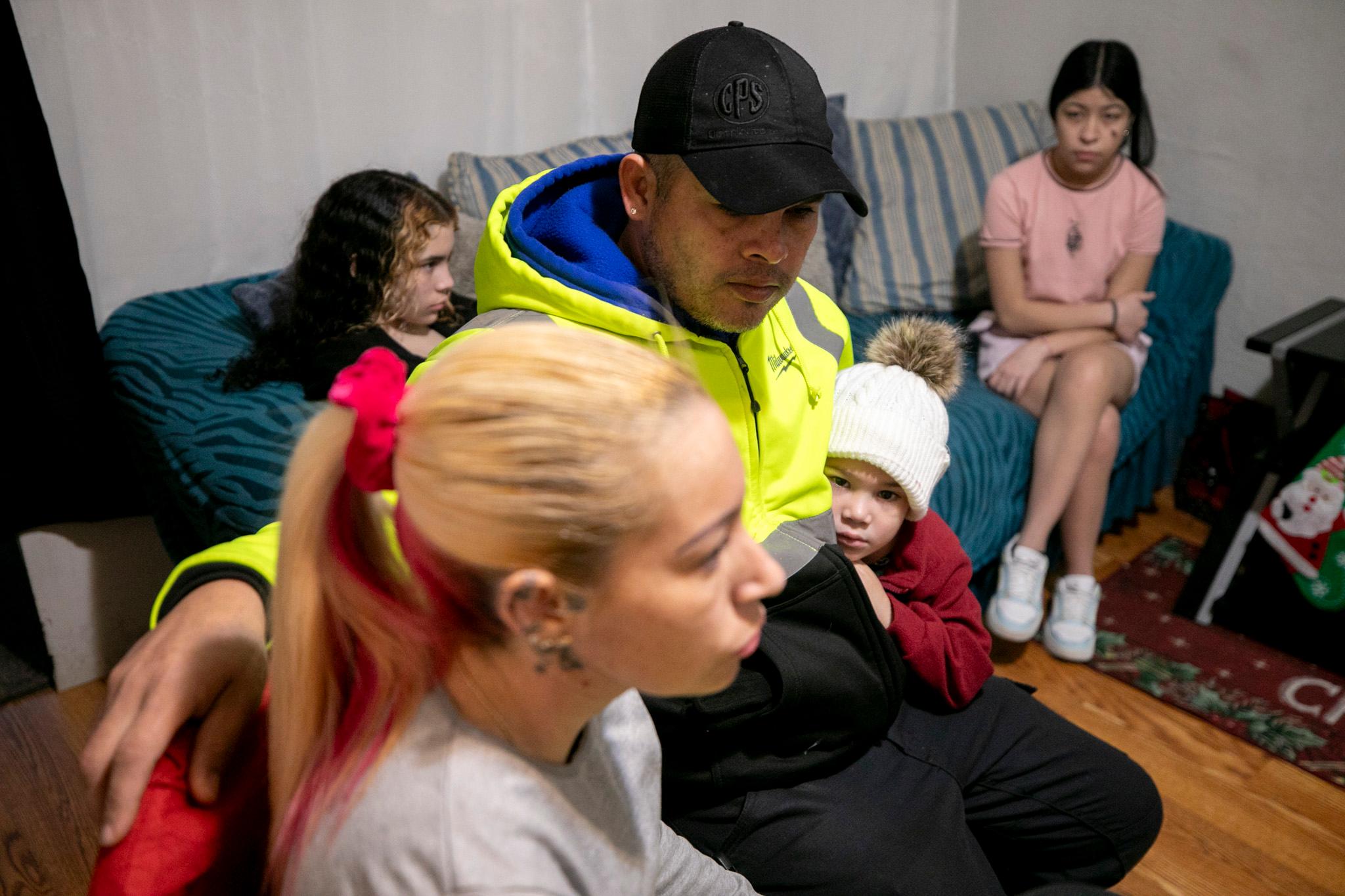 A family, with a woman in the foreground and a man and children behind her, sit on a two sofas in a small apartment.