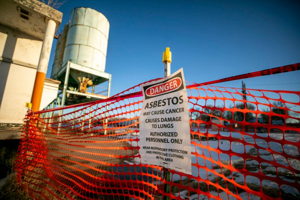 A sign reading asbestos sits on an orange fence with a large white grain silo in the background.