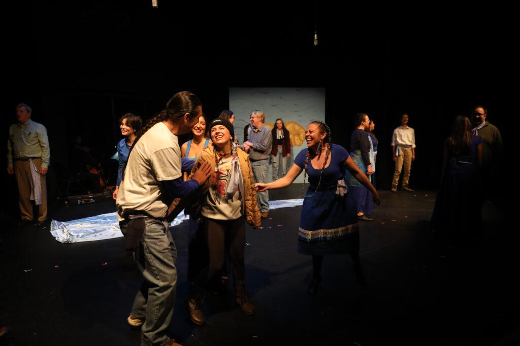 A stage performance featuring a group of actors with one actor, wearing a brown jacket, center, interacting enthusiastically with others. The background is dark with a simple set.