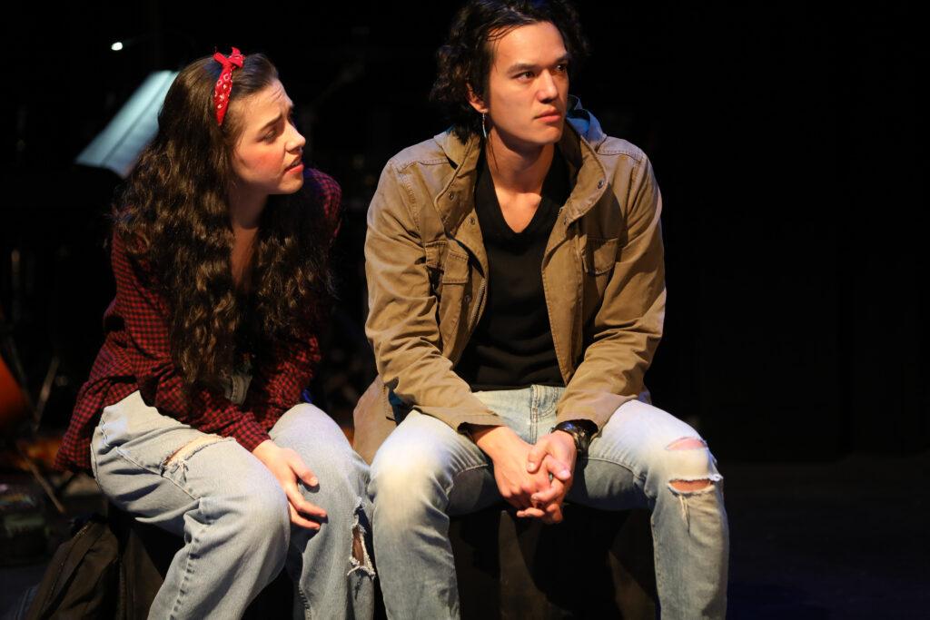 A woman with long dark hair and a red headband sits next to a man with wavy hair on stage in a dramatic moment.