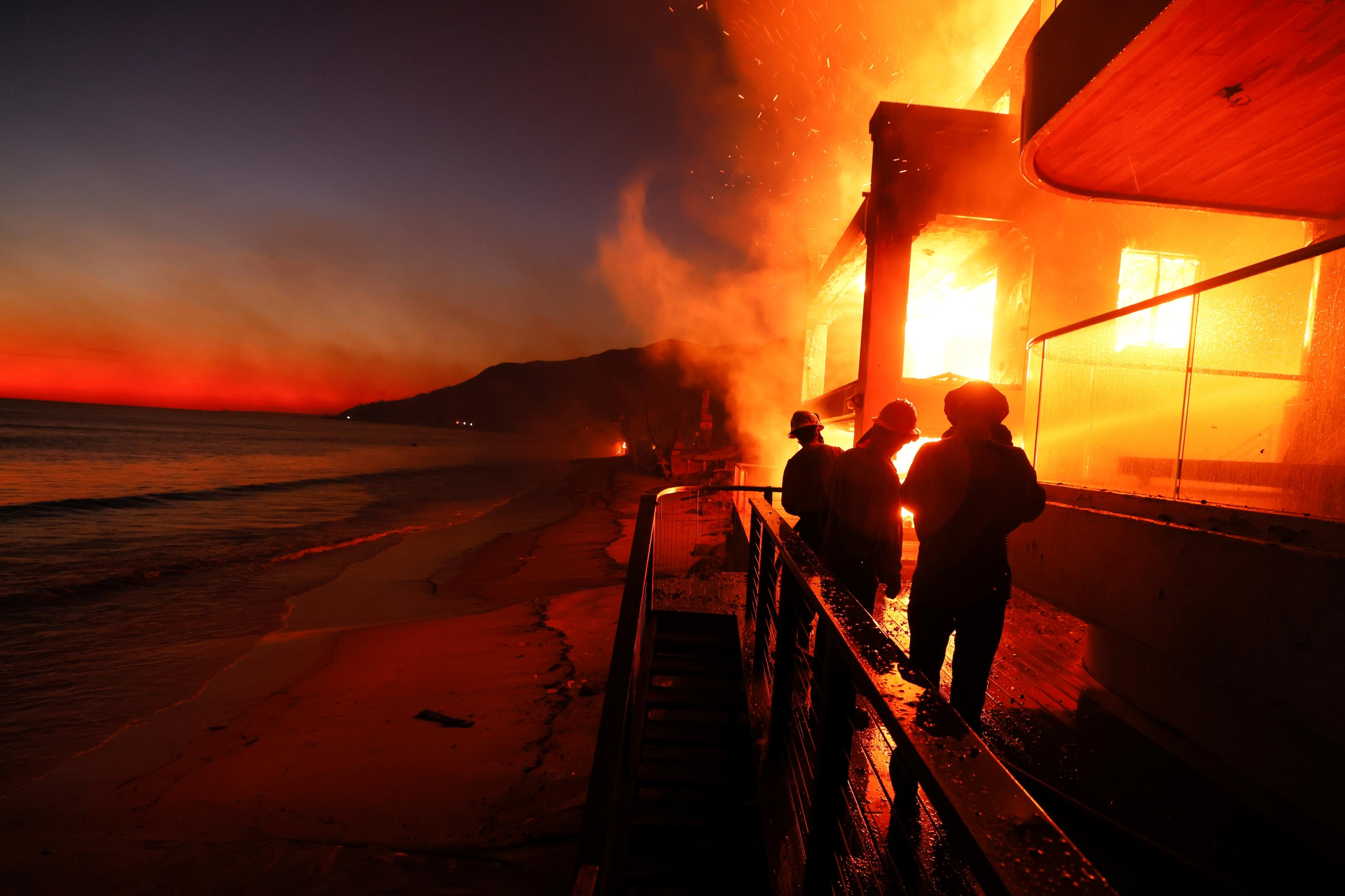 Firefighters stand in the glow of flames battling the fire of a beach front house in California