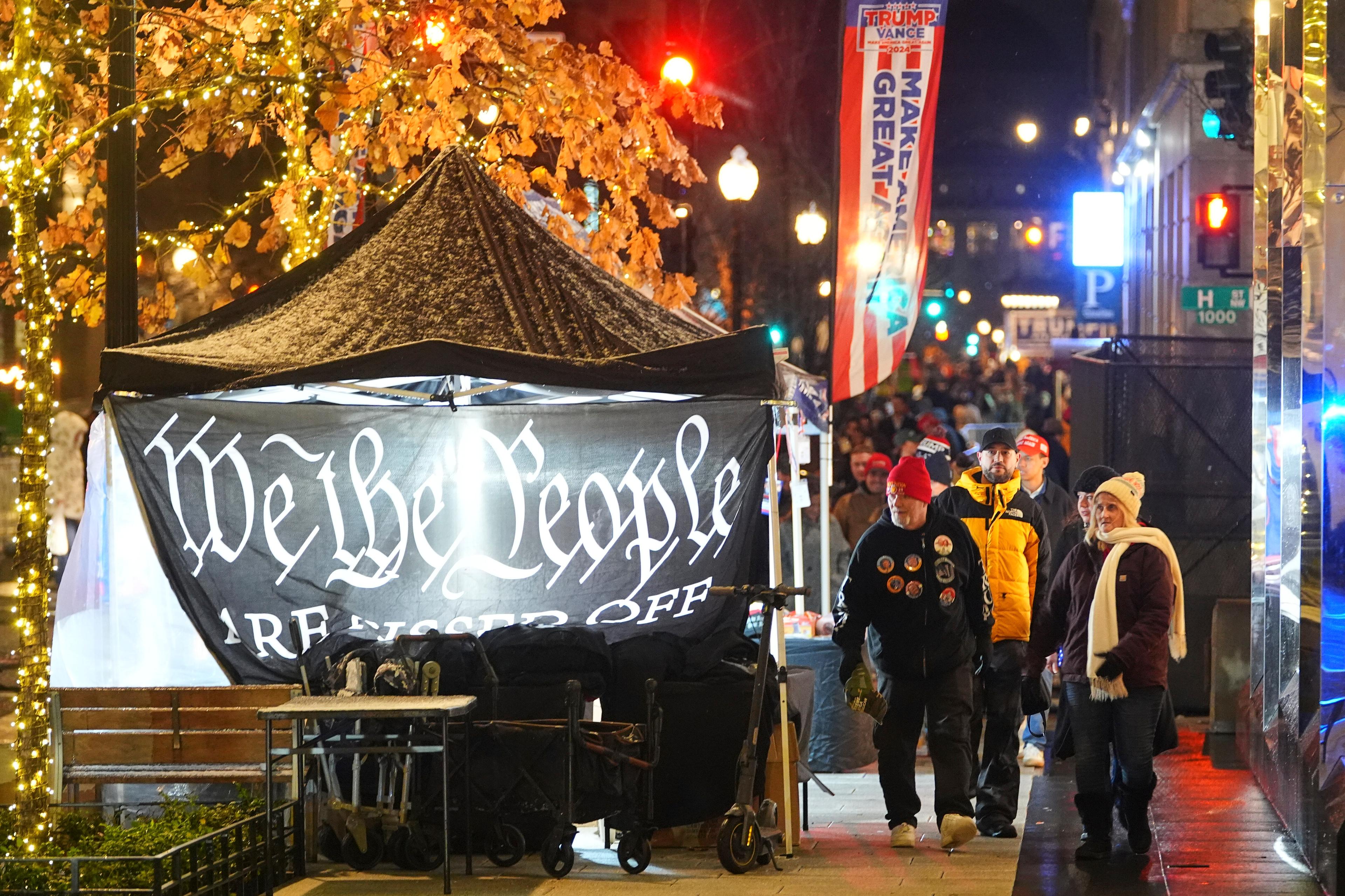 A night scene of a tent with a We The People flag as a crowd walks past