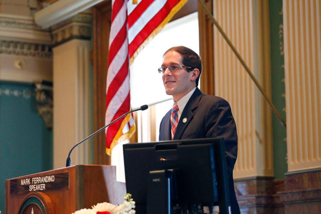 A man in a suit glasses stands at a lecturn at the front of the House chamber