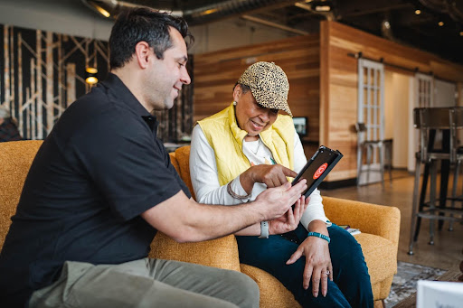 A man is seated next to a woman and is holding an iPhone to show her how it works.