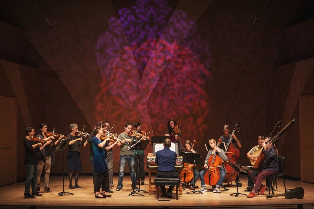 Musicians from the Baroque Chamber Orchestra of Colorado play passionately on stage, accompanied by a striking large projection.