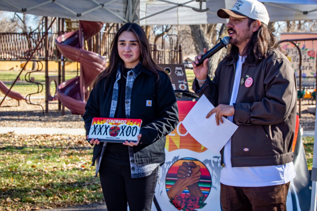 a woman holds a license plate, a man speaks into a microphone at a park