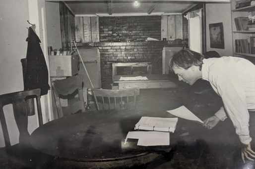 A black and white photo shows a man leaning over a table looking at documents inside a historic house in Antonito, CO.