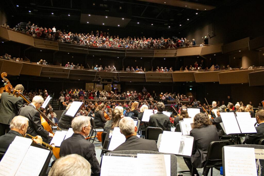 A vibrant concert scene featuring the Grand Junction Symphony Orchestra performing, with an engaged audience in attendance.