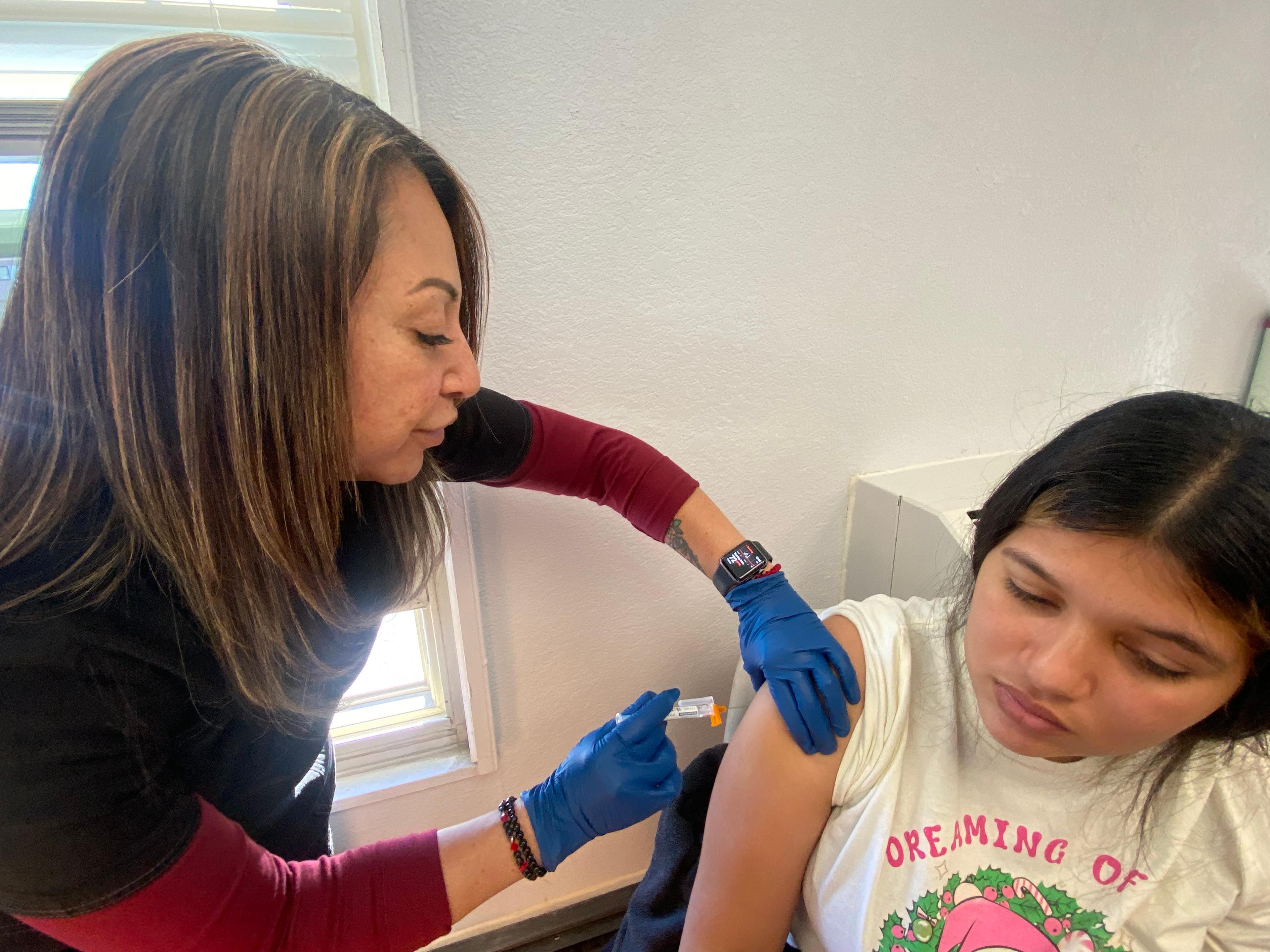 A medical professional wearing black scrubs over a red long-sleeved shirt and blue gloves administers a vaccine to the shoulder of a younger woman.