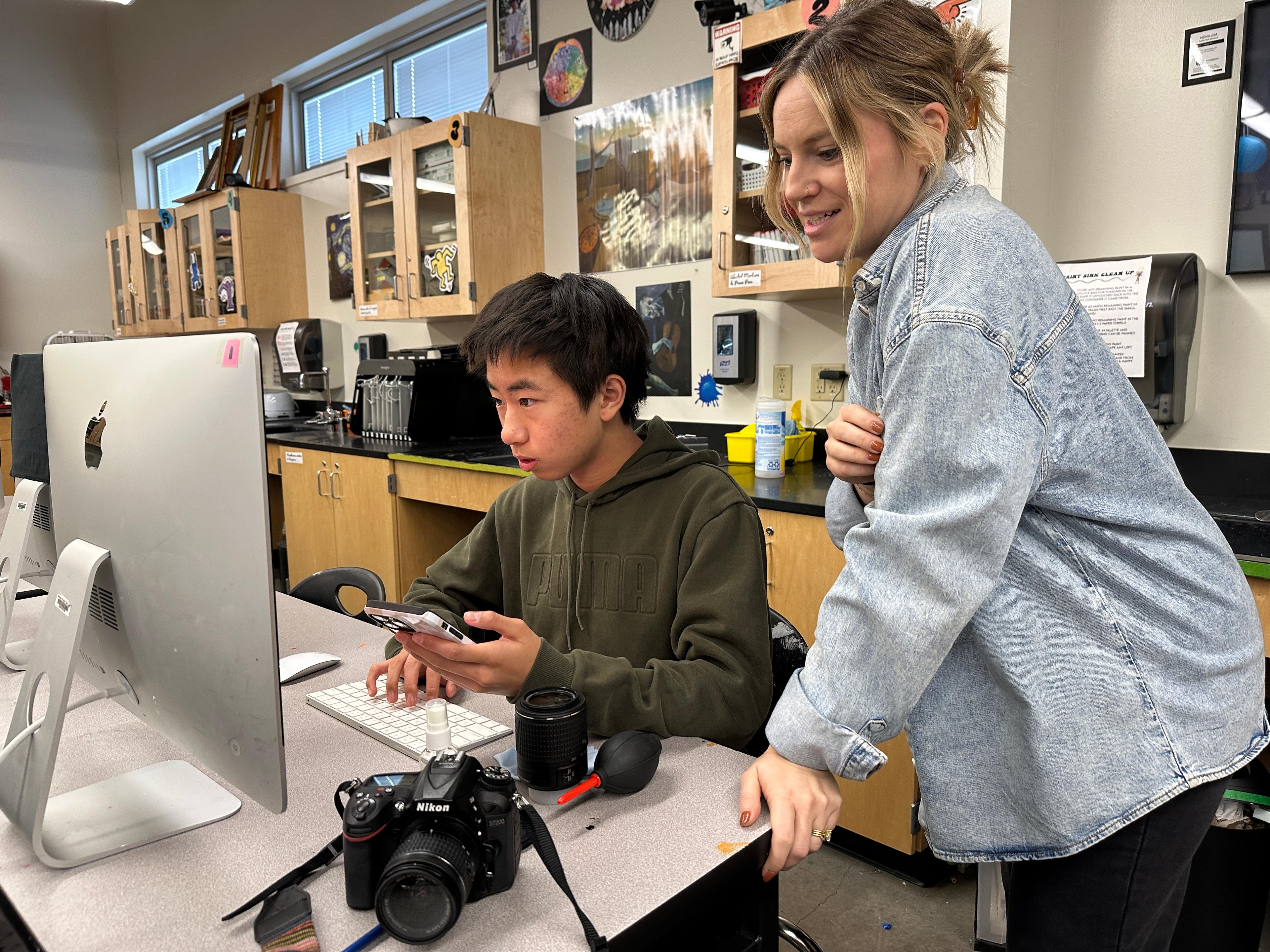 A teacher in a light demin shirt leans on a desk where a student is typing on a computer while holding a cell phone in his left hand.