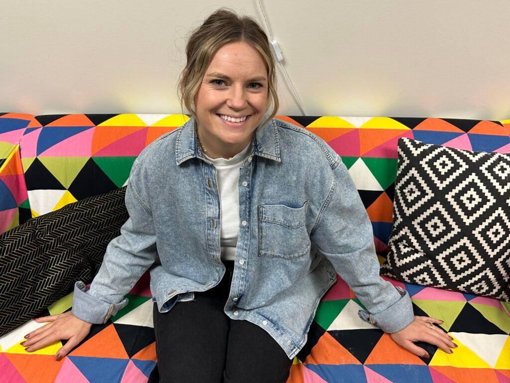 A woman wearing a light demin shirt sits with her hands out on a couch. The couch has a geometric design of colorful triangles.