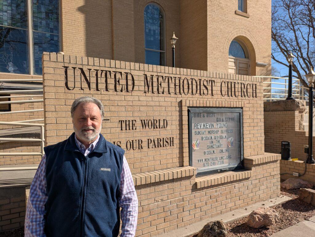 Rev. Kevin Young, wearing a checkered dress shirt and a blue vest, stands in front of the United Methodist Church in Montrose.