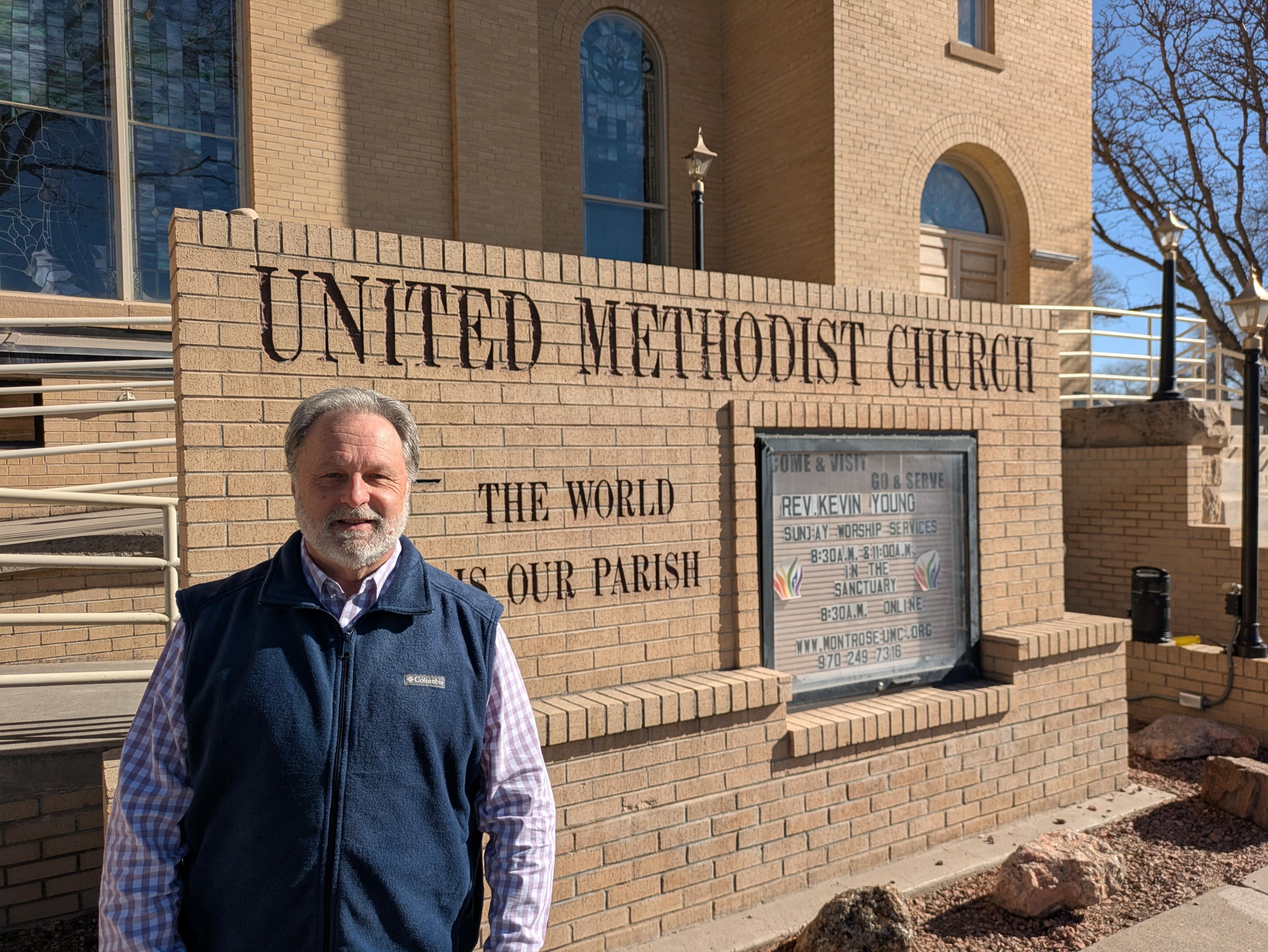 Rev. Kevin Young, wearing a checkered dress shirt and a blue vest, stands in front of the United Methodist Church in Montrose.