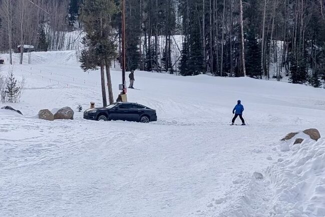 A black sedan sits at the base of a ski run with skiers in the background.