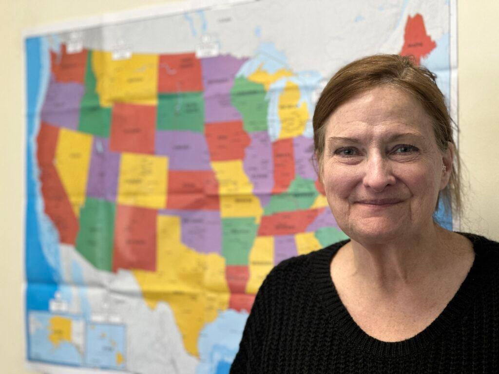 A closeup portrait of an older woman with reddish hair smiling. Her face is in the right foreground and a colorful map of the U.S. is in the background