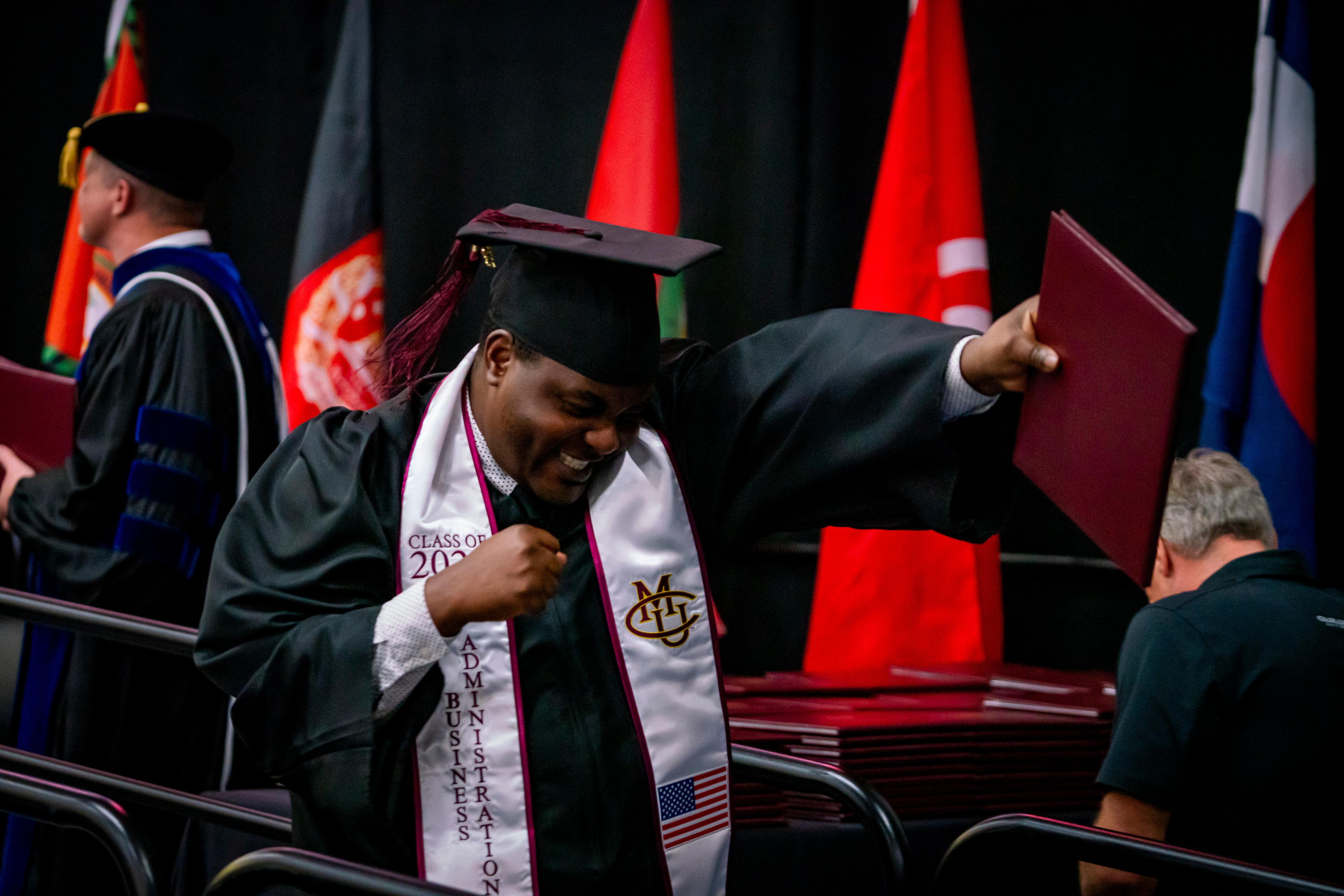 Mathias Mulumba, wearing a black cap and gown for graduation with a white Colorado Mesa University stole, pumps his fist while crossing the stage during CMU's commencement ceremony in Grand Junciton.