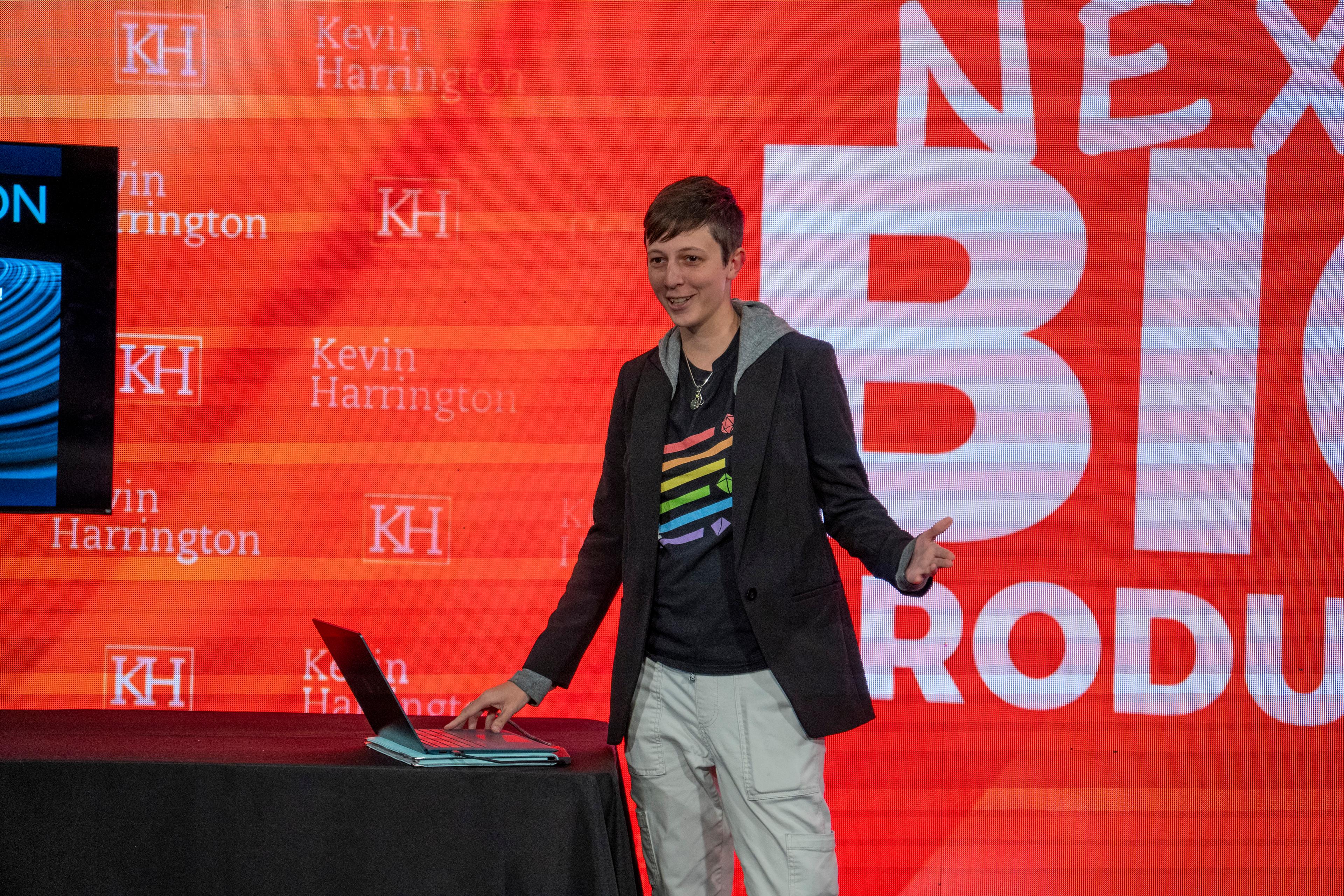 A woman with short hair standing in front of orange sign with a table in front of her on which her hand is on the keyboard.