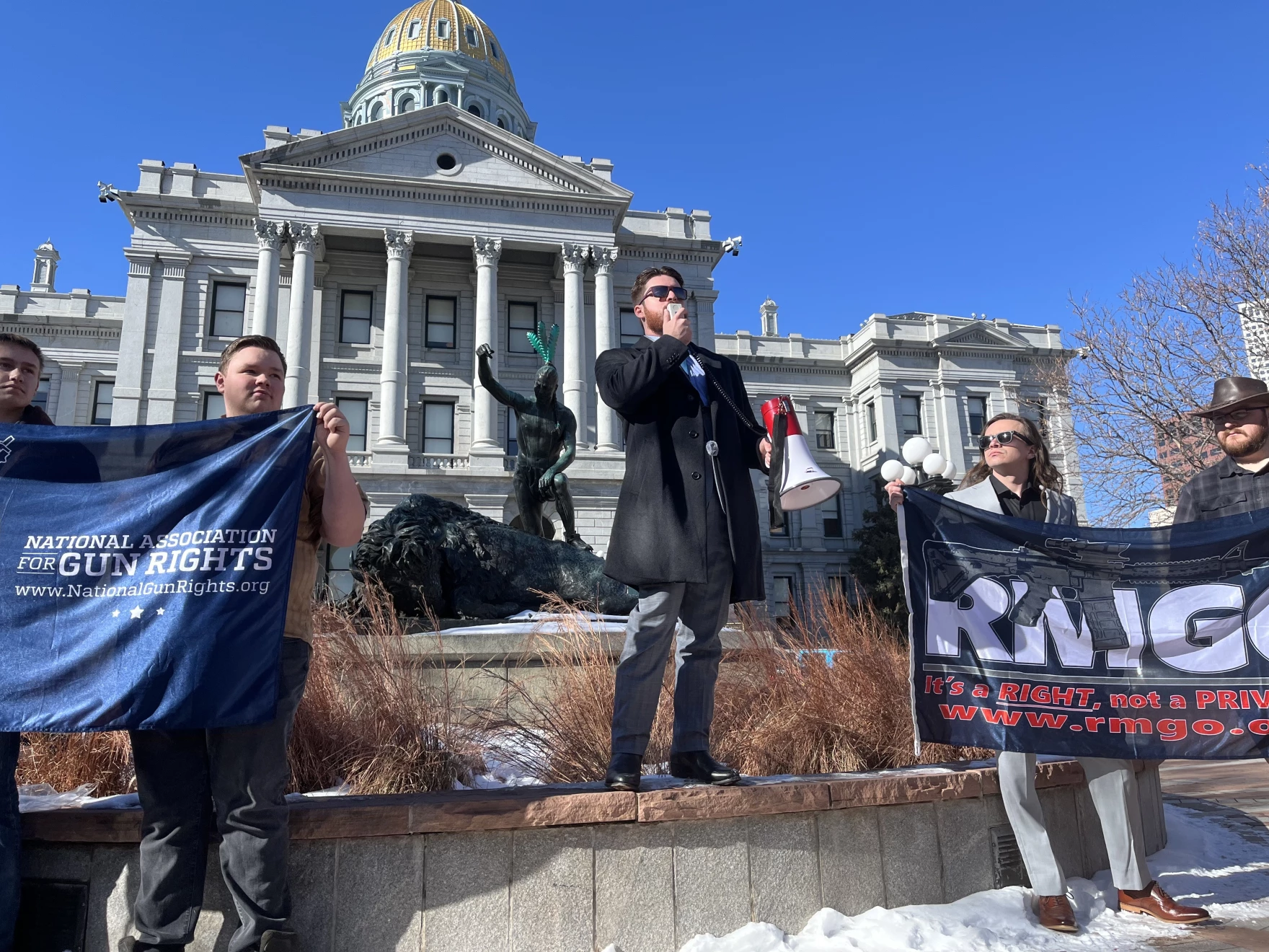 A man with a megaphone stands in front of the Capitol with people holding flags on either side of him.