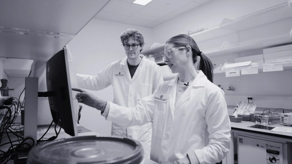 A man and a woman stand in a lab in white lab coats staring at testing equipment