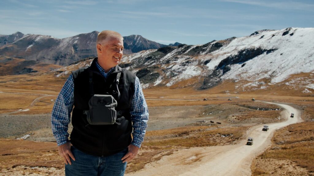 Terry Meyers stands on a dirt road with mountains and ATVs in the background