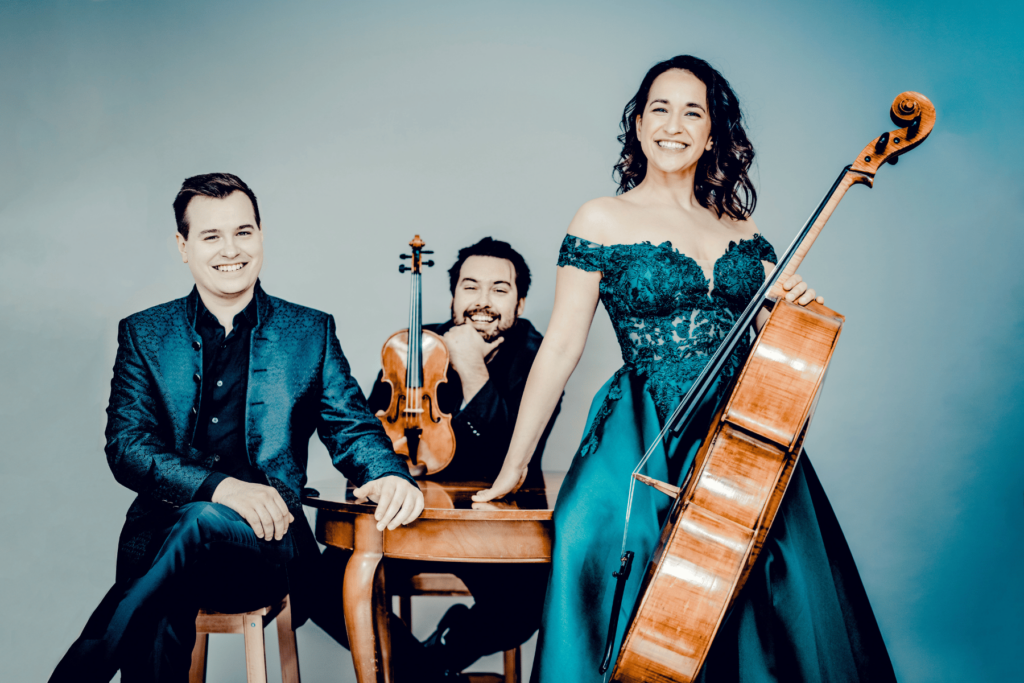 Three musicians in formal wear pose with some instruments for a photo against a turquoise backdrop, for their upcoming concert at the Vilar Performing Arts Center.