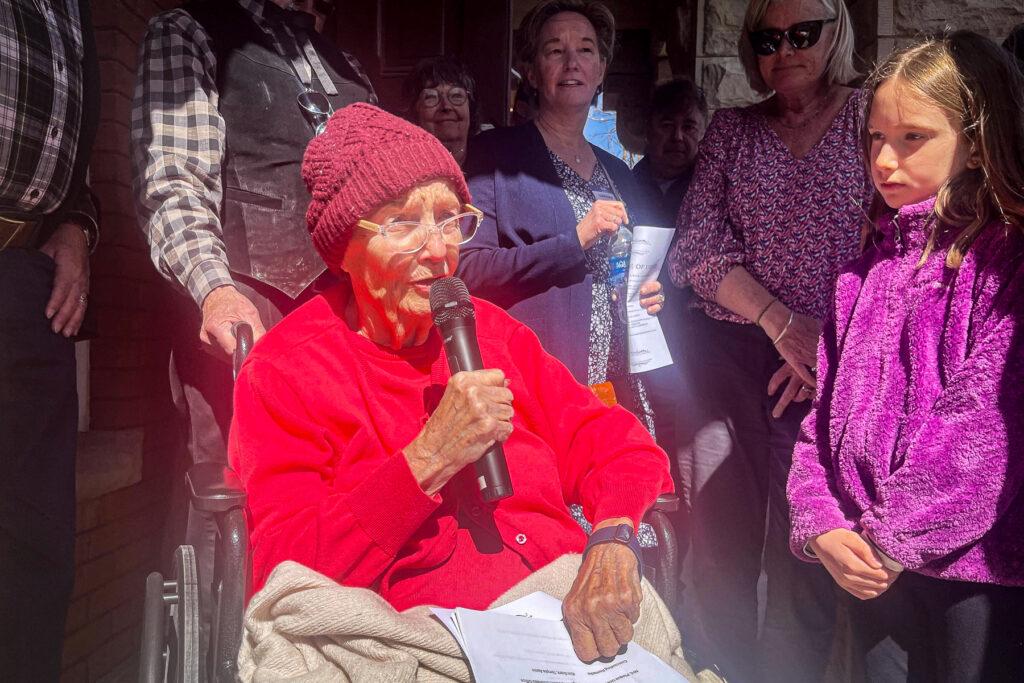 A photo of Dana Crawford in a red sweater and beanie holding a microphone in a wheelchair