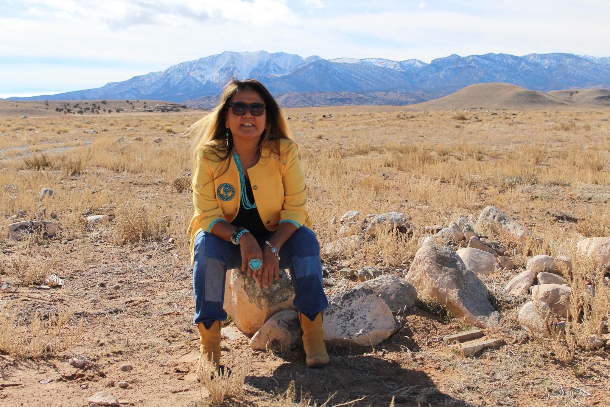 native american middle-aged woman dressed in jeans, cowboy boots and turquoise jewelry sitting on rock in desert in front of mountains