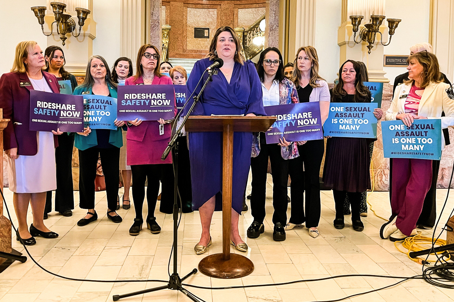 Rep. Jenny Wilford stands at a lectern in the capitol foyer, with fellow lawmakers standing around her holding signs supporting the bill