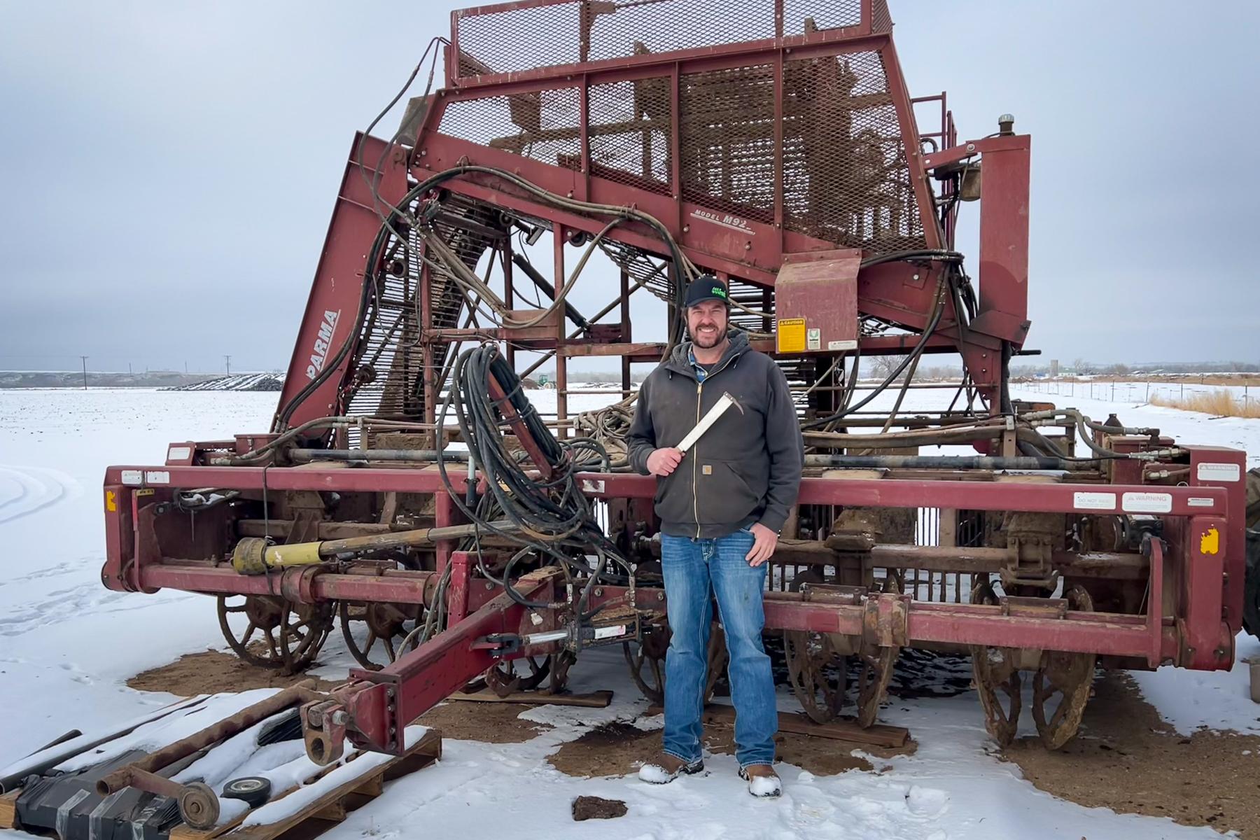 A man who is a sugar beet farmer stands in front of a large, red sugar beet harvester. He's holding a special knife with a curved tip that farmworkers used to use to harvest sugar beets by hand.