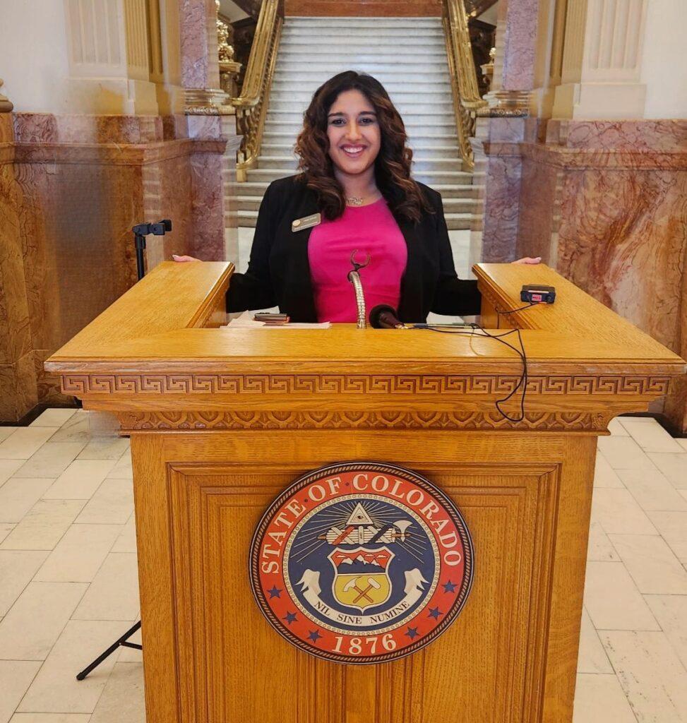 woman with curly hair standing at wooden podium that has state of Colorado seal