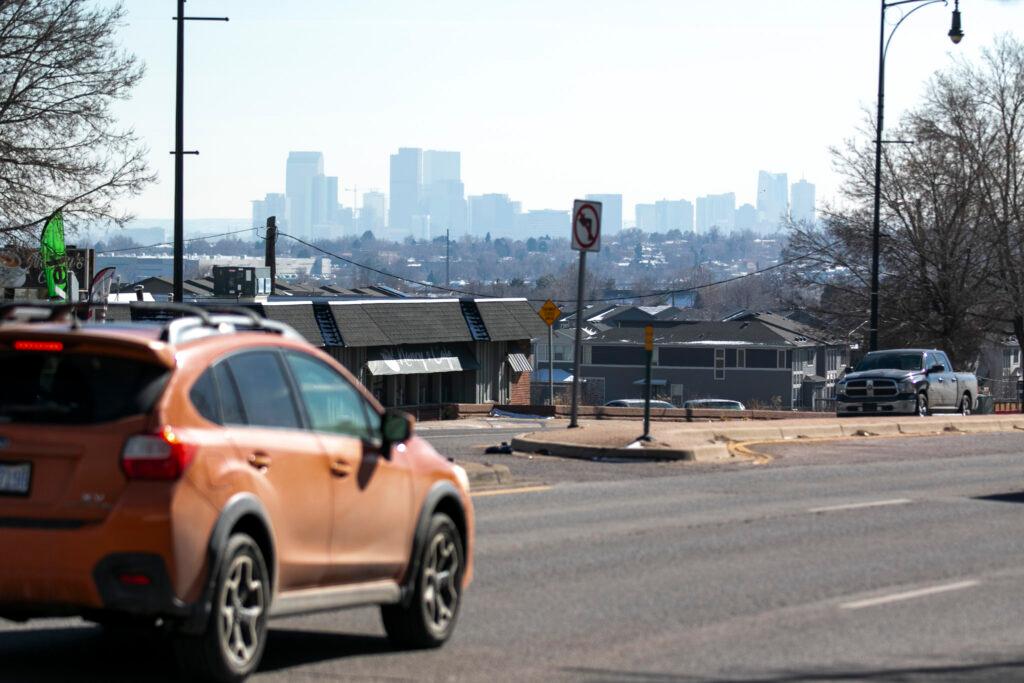 Downtown Denver sits amid haze as an orange car passes by on a road.