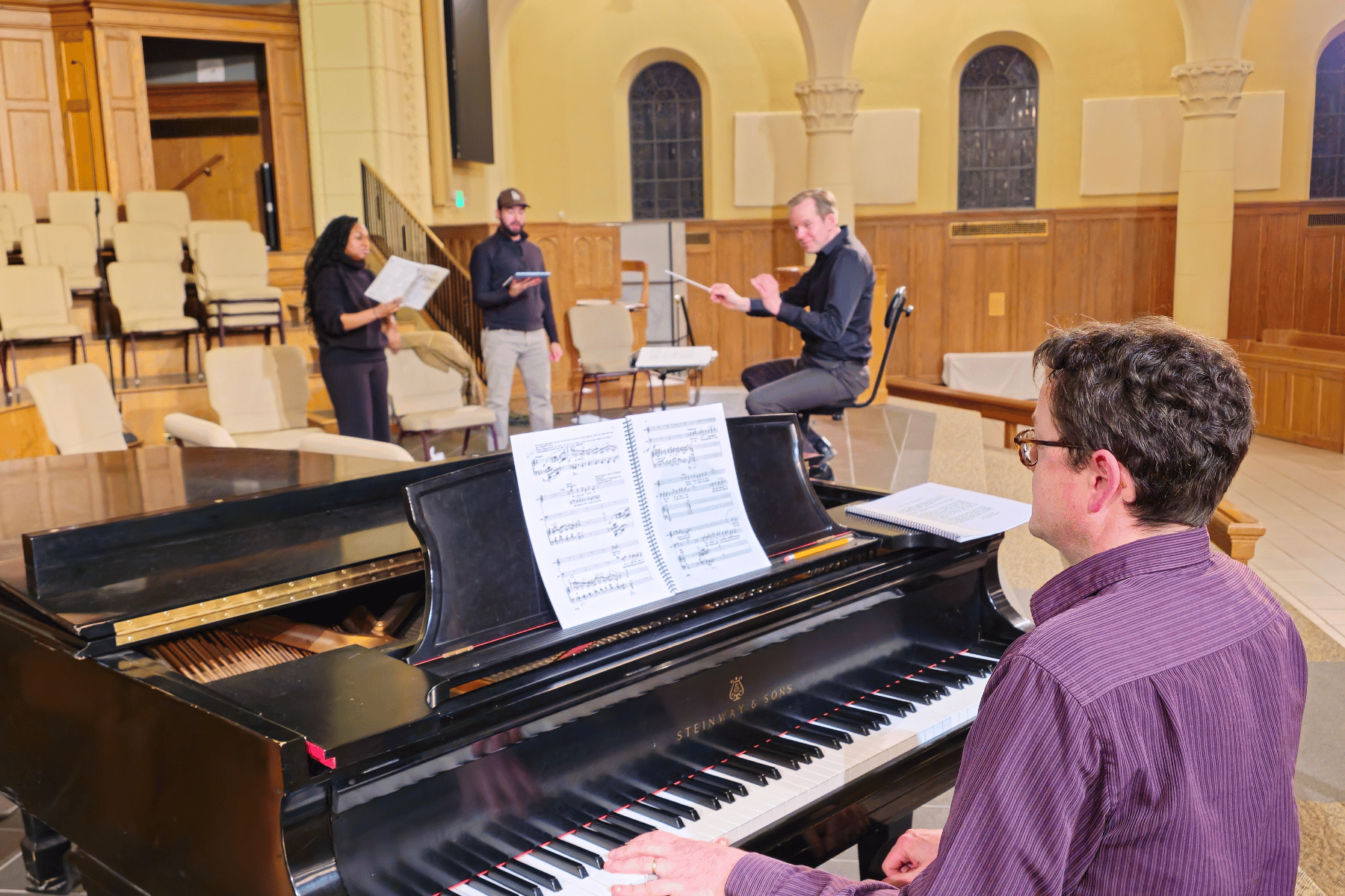 A man plays piano in a church, collaborating with a conductor and singers during a rehearsal for All About Love.