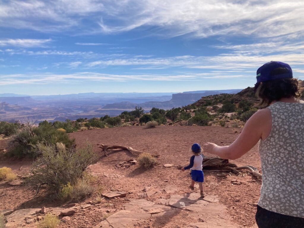 A boy walks outside on red rocks. In teh background is a vast view of mesas of southern Colorado.