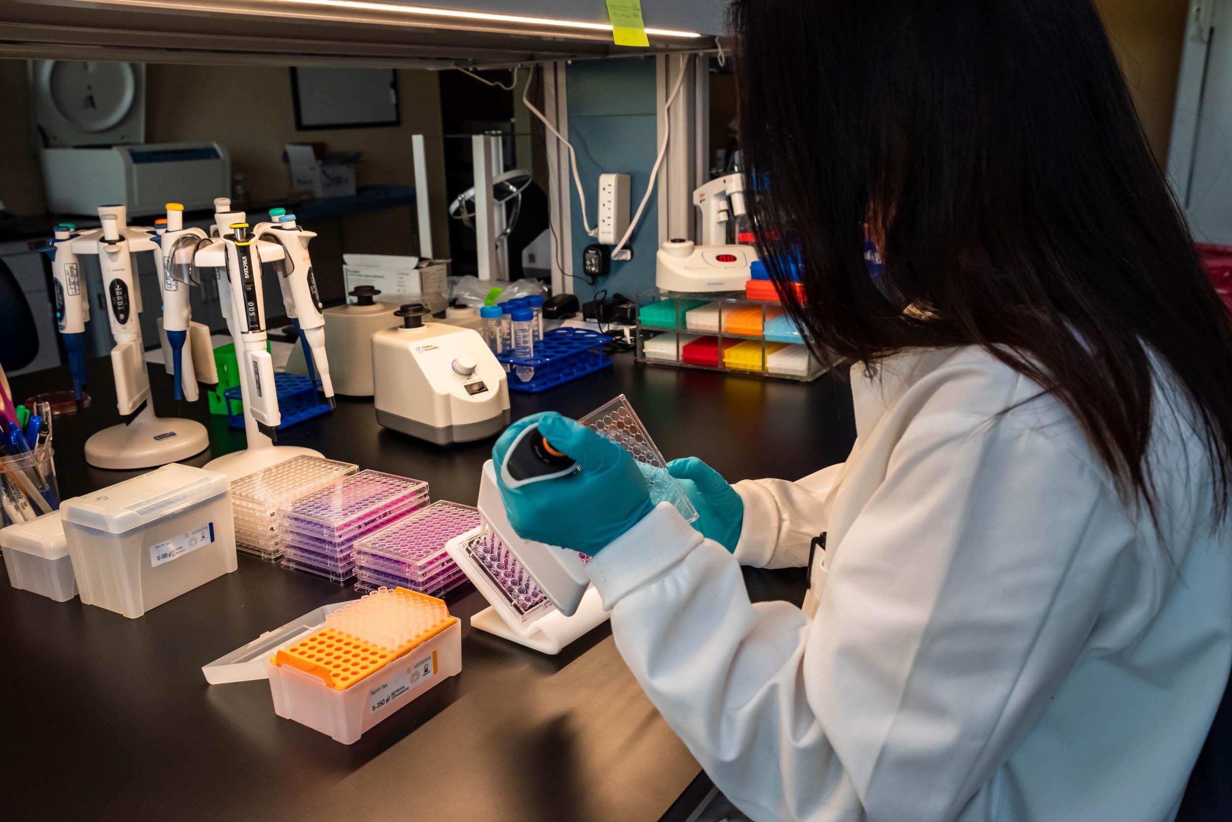 A woman, whose face is obscurred by her long black hair, prepares vials in a science lab. She is wearing a white coat and blue medical gloves.